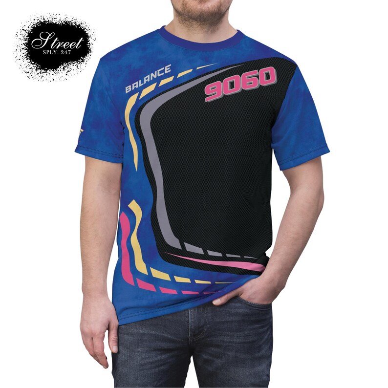 Puede incluir: Una camiseta azul de manga corta con un dise&ntilde;o gr&aacute;fico negro, gris, rosa y amarillo. El dise&ntilde;o gr&aacute;fico presenta la palabra "BALANCE" y el n&uacute;mero "9060".