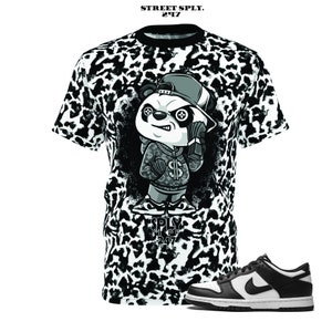May include: A black and white t-shirt with a panda bear wearing a hat and holding a phone. The panda bear has a black eye patch and a chain around its neck. The t-shirt has a leopard print pattern. The t-shirt is paired with black and white sneakers.