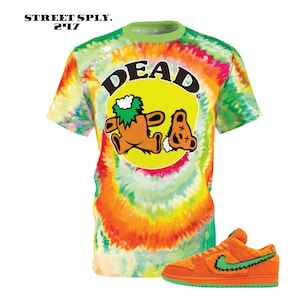 May include: A tie-dye t-shirt featuring a yellow circle with the text "DEAD" in black. Inside the circle are two cartoon bears, one orange and one brown. The bears are wearing green hats and have a green background. The t-shirt is paired with orange and green trainers.