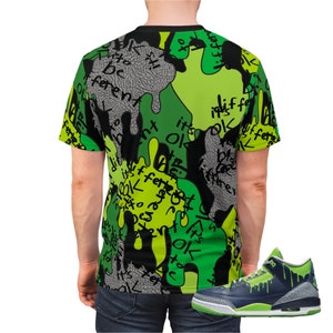 Puede incluir: Una camiseta negra, verde y gris con un estampado de camuflaje y las palabras "it's ok to be different" impresas en ella. Tambi&eacute;n se muestra un par de zapatillas verdes y azules con un dise&ntilde;o de goteo.