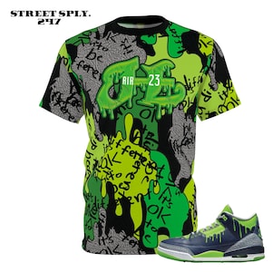 Puede incluir: Una camiseta negra con un estampado de graffiti verde y amarillo con el texto "AIR 23". La camiseta se combina con una zapatilla azul marino y verde con suela verde.