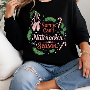 May include: Black sweatshirt with a festive design. The graphic includes pink ballet shoes, candy canes, and the text "Sorry, Can't, Nutcracker, Season." The design is surrounded by green sprigs and white stars.