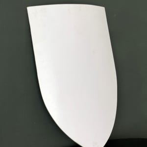 May include: A white, shield-shaped object with a rounded top and a pointed bottom. The object is smooth and appears to be made of a solid material. The background is a dark grey colour, and the object is reflected on a black surface.