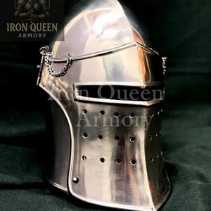 Puede incluir: Un yelmo de caballero plateado pulido con visera y detalle de cadena. El yelmo tiene una parte superior redondeada y una serie de pequeños agujeros en la parte delantera inferior. El logotipo "Iron Queen Armory" es visible en la esquina superior izquierda.