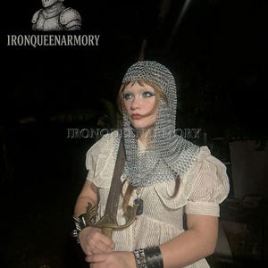 May include: A person wearing a chainmail coif and holding a sword. The person is wearing a cream-coloured blouse with short, puffed sleeves and a striped pattern. The image includes the text "IRONQUEENARMORY" and a logo of a knight's helmet.