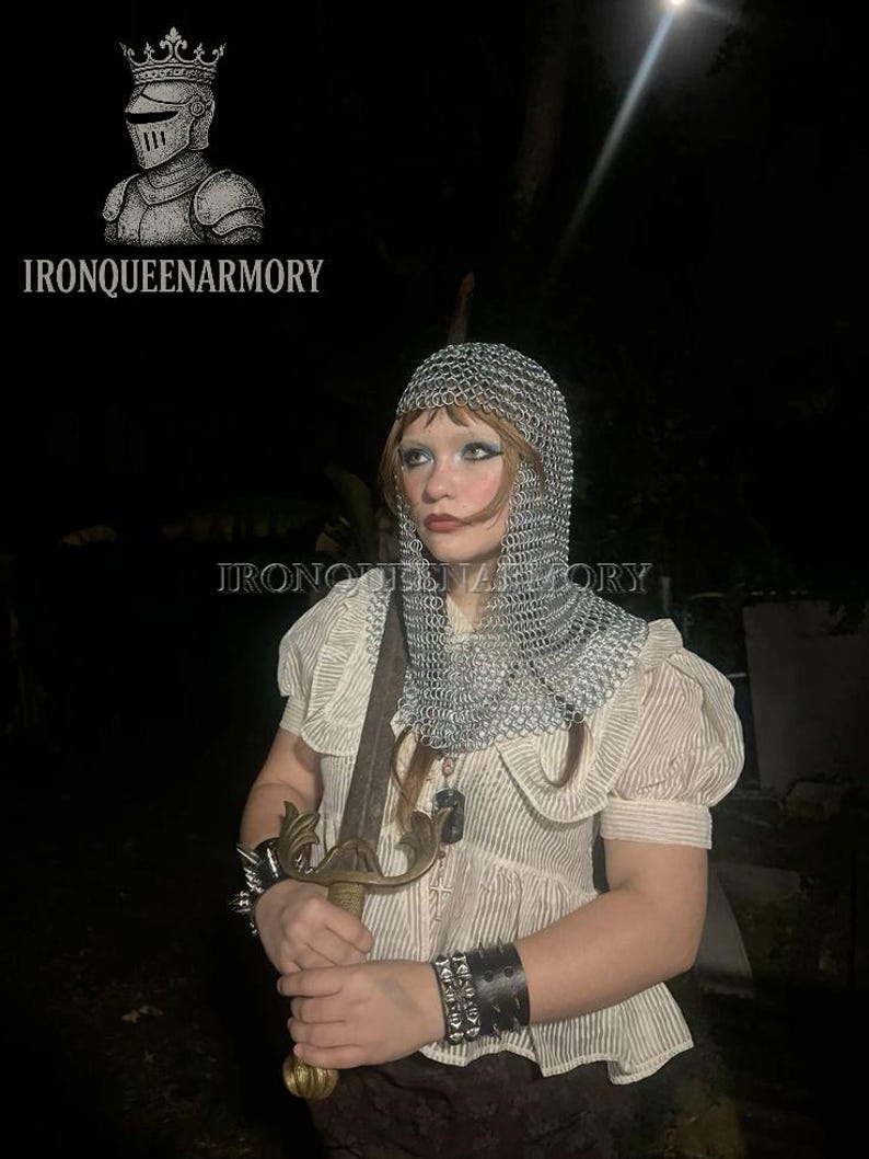 May include: A person wearing a chainmail coif and holding a sword. The person is wearing a cream-coloured blouse with short, puffed sleeves and a striped pattern. The image includes the text "IRONQUEENARMORY" and a logo of a knight's helmet.