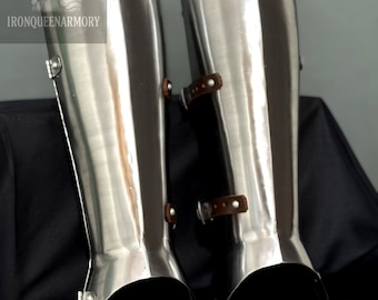 May include: Pair of polished steel greaves, part of medieval armor. The leg guards have a shiny, reflective surface and are secured with leather straps and buckles. The image includes the text "IRONQUEENARMORY".
