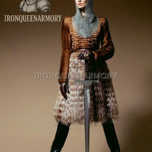 May include: A person in a medieval-inspired outfit, holding a sword. They wear a chainmail hood, a brown fur coat, and a fur skirt. The outfit is completed with black leather boots and gloves. The image includes the text "IRONQUEENARMORY".