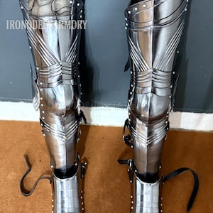 May include: Pair of polished steel leg armor with riveted accents and leather straps. The armor features articulated segments and a pointed lower section. The text "IRONQUEEN ARMORY" is visible on the left leg armor.