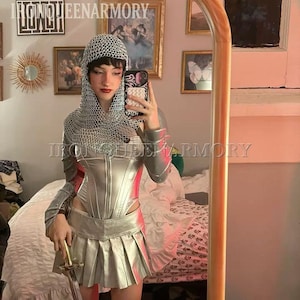 May include: A silver-coloured armour set, including a corset, pleated skirt, and chainmail hood. The outfit is paired with matching boots and a sword. The text "IRONQUEENARMORY" is visible. A mirror reflects the outfit.