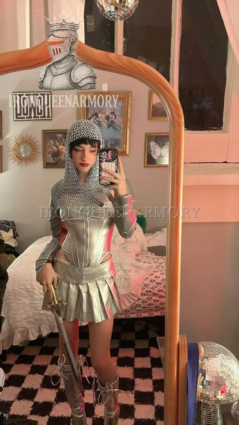 May include: A silver-coloured armour set, including a corset, pleated skirt, and chainmail hood. The outfit is paired with matching boots and a sword. The text "IRONQUEENARMORY" is visible. A mirror reflects the outfit.
