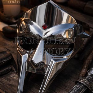 May include: A highly polished silver metal mask with a red gem and spikes. The mask has a reflective surface and is displayed on a wooden surface. The text "IRON QUEEN ARMORY" is visible on the mask.