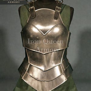 May include: A silver breastplate with a layered design, displayed on a black mannequin. The armour has leather straps and buckles. The text "Iron Queen Armory" is visible on the armour.