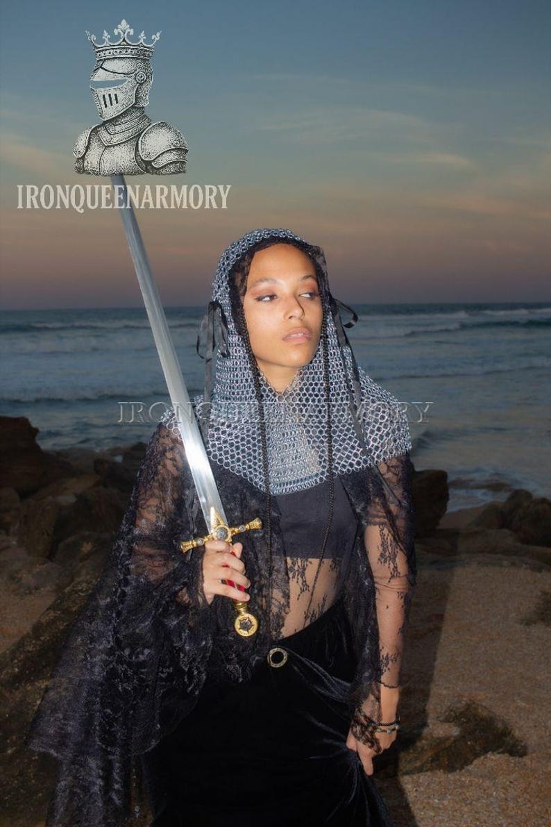 May include: A person holding a long sword with a gold hilt and a silver blade. The person is wearing a chainmail hood and a black lace cape. The image includes the text "IRONQUEENARMORY".
