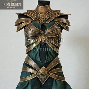 May include: A detailed shot of a fantasy costume. The armor is a metallic gold color with green accents and gemstones. The costume is displayed on a mannequin and features a breastplate, shoulder armor, and a belt. The dress underneath is dark green.