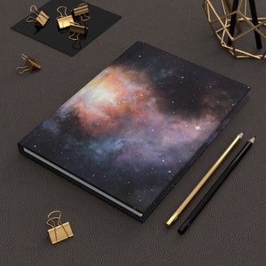 Op de afbeelding: Een zwart notitieboek met een galaxy-print op de omslag. De omslag heeft een nevel met tinten oranje, roze en paars. Het notitieboek ligt op een donkergrijze ondergrond met twee potloden en gouden paperclips.