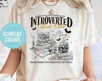 May include: A white t-shirt featuring a black and gold graphic design that reads "Proudly Anti-Social Member of the Introverted Book Club" with a skull and stack of books. The text below the graphic says "Making Friends with Characters, Not People".