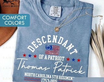 May include: A blue jean Comfort Colors t-shirt with the text "Descendant of a Patriot Thomas Patrick" and an American flag design. The shirt also includes the text "North Carolina 5th Regiment 1753-1815". A gold necklace rests on the shirt.