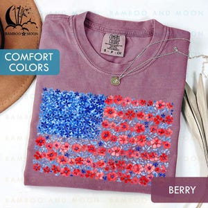 May include: Berry-colored t-shirt with a floral American flag design. The flag is composed of blue and red flowers. The shirt has the words "COMFORT COLORS" printed on it.