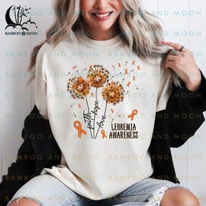 Peut inclure: Sweat-shirt blanc avec un ruban orange et un motif de pissenlit. Le texte "Foi, Espoir, Amour" et "Sensibilisation à la leucémie" est imprimé sur le t-shirt.