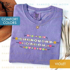 May include: A violet Comfort Colors t-shirt featuring the words "PEDIATRIC NURSE" in a decorative font, embellished with hearts, flowers, and smiley faces. A gold necklace is draped across the shirt.