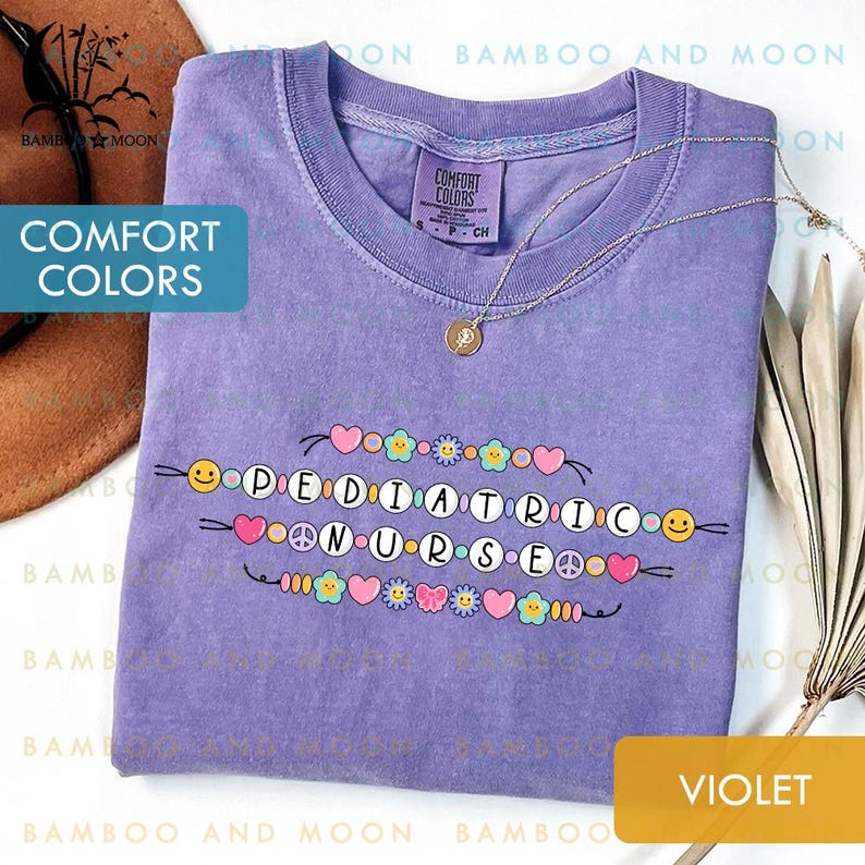 May include: A violet Comfort Colors t-shirt featuring the words "PEDIATRIC NURSE" in a decorative font, embellished with hearts, flowers, and smiley faces. A gold necklace is draped across the shirt.