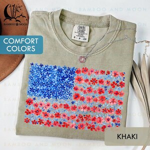 May include: Khaki-colored t-shirt with a floral American flag design. The flag is composed of blue and red flowers. The shirt has the words "Comfort Colors" printed on it. A silver necklace with a pendant is draped across the shirt.