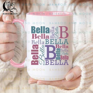 May include: White ceramic mug with a pink interior and handle. The mug features the name "Bella" in various fonts and colours, including teal, purple, and pink. The mug is held by a person.