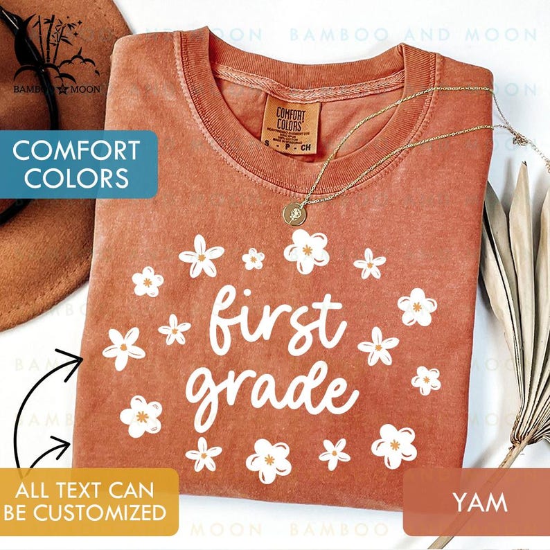 May include: A terracotta-colored t-shirt with the words "first grade" in white script, surrounded by white flower graphics. The shirt has a crew neck and short sleeves. The text "Comfort Colors" is visible on the tag. A gold necklace with a pendant is on the shirt.