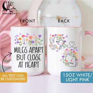 May include: White ceramic mug with a pink handle and interior, featuring floral designs and the text "Miles Apart But Close At Heart." The back of the mug shows state outlines with names and floral accents. The mug holds 15 oz.