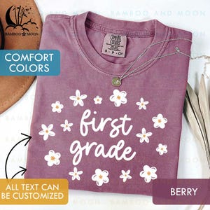 May include: Berry-colored t-shirt with the words "first grade" in white, surrounded by white flowers with yellow centers. The shirt has a crew neck and short sleeves. The text "COMFORT COLORS" is visible on the tag. A silver necklace with a pendant is draped on the shirt.