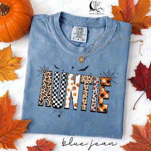 May include: A blue short-sleeved t-shirt with a crew neckline and a Comfort Colors brand label. The front features the word AUNTIE in block letters with various patterns including leopard print, black and white checkers, pumpkins, and orange plaid.