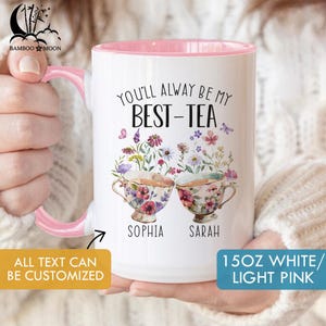 May include: White ceramic mug with a pink interior and handle, featuring the text "YOU'LL ALWAYS BE MY BEST-TEA." Two teacups with floral designs are below the text, with the names "SOPHIA" and "SARAH" beneath each. The mug is 15oz and the text "ALL TEXT CAN BE CUSTOMIZED" is also visible.