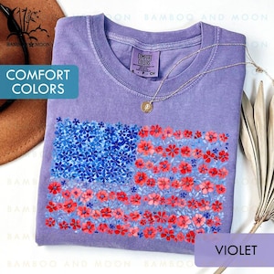 May include: A violet-colored t-shirt with a floral American flag design. The flag is composed of blue and red flowers. The shirt has the words "COMFORT COLORS" printed on it. A gold necklace is draped on the shirt.