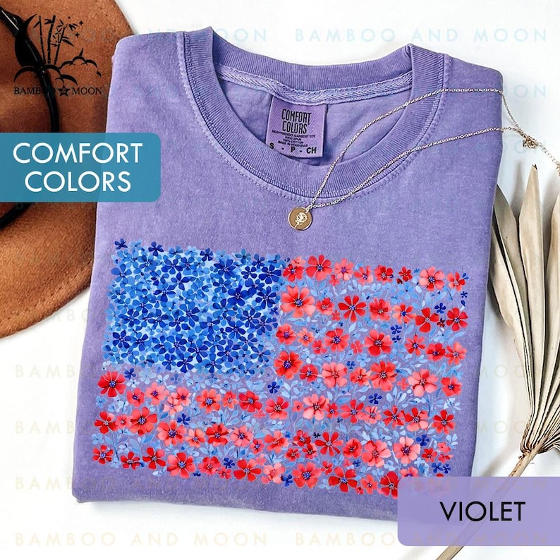 May include: A violet-colored t-shirt with a floral American flag design. The flag is composed of blue and red flowers. The shirt has the words "COMFORT COLORS" printed on it. A gold necklace is draped on the shirt.