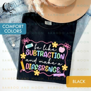 Pode incluir: Camiseta preta com a frase "be like SUBTRACTION and make a DIFFERENCE" em um design colorido com gráficos escolares. A camiseta é mostrada com jeans e um chapéu. Feita de material Comfort Colors.