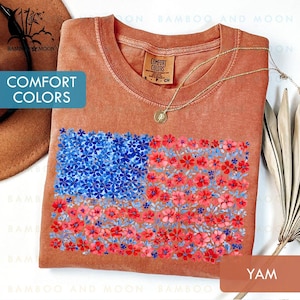 May include: A yam-colored t-shirt with a floral American flag design. The flag features blue flowers in the upper left corner and red flowers in the remaining area. The shirt has the words "COMFORT COLORS" on the tag.