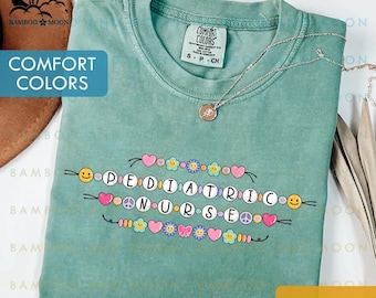 May include: Seafoam green t-shirt with the words "PEDIATRIC NURSE" spelled out with decorative beads, hearts, flowers, and smiley faces. The shirt has a crew neck and short sleeves. A silver necklace with a pendant is draped on the shirt.