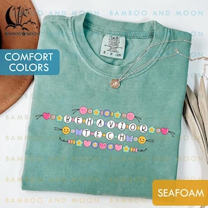 May include: Seafoam green t-shirt with the words "BEHAVIOR TECH" in a decorative font, adorned with hearts, flowers, and smiley faces. The shirt is labeled "Comfort Colors" and has a small necklace draped across it.