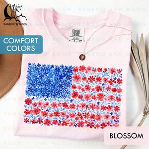 May include: A light pink Comfort Colors t-shirt featuring a floral American flag design. The flag is composed of blue flowers in the upper left corner and red flowers in the stripes. The shirt has the text "BLOSSOM".
