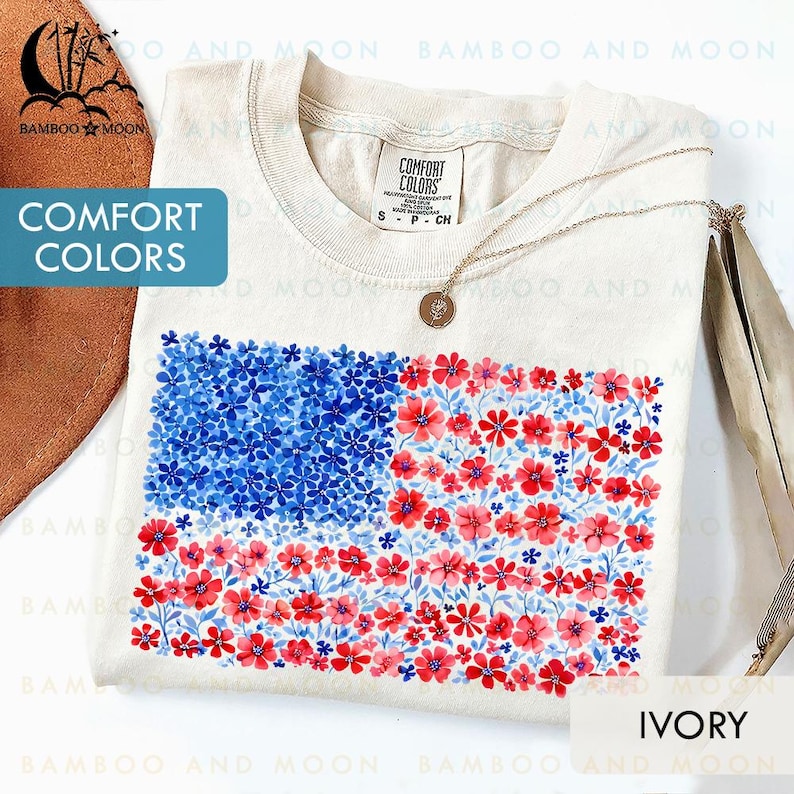 May include: Ivory-colored t-shirt featuring a floral American flag design. The flag's stars are represented by blue flowers, and the stripes are red flowers. The shirt has a crew neck and short sleeves. The text "COMFORT COLORS" is visible.