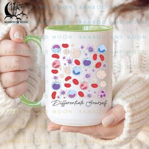 May include: White ceramic mug with a green handle and rim, featuring colorful illustrations of cells and the text "Differentiate Yourself." The mug is held by a person wearing a white sweater.