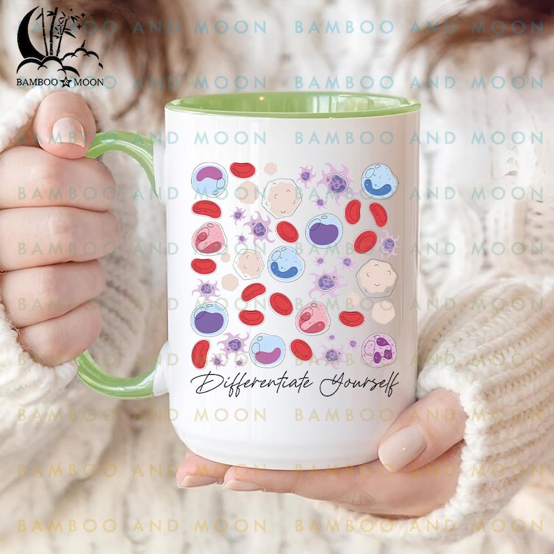 May include: White ceramic mug with a green handle and rim, featuring colorful illustrations of cells and the text "Differentiate Yourself." The mug is held by a person wearing a white sweater.