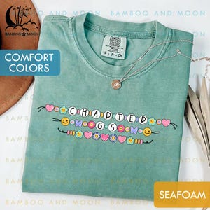 Pode incluir: Camiseta Comfort Colors verde-mar com o texto "CHAPTER 6.5" em uma fonte decorativa, adornada com corações coloridos, flores e rostos sorridentes. Um colar de prata com um pingente repousa sobre a camisa.