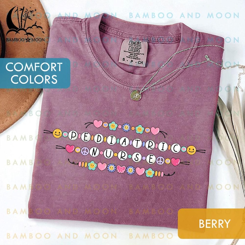 May include: Berry-coloured t-shirt featuring the text "PEDIATRIC NURSE" in a decorative font, embellished with hearts, flowers, peace signs, and smiley faces. A silver necklace with a pendant rests on the shirt. The shirt is labelled "Comfort Colors".