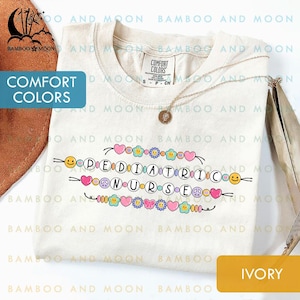 May include: Cream-coloured t-shirt featuring the text "PEDIATRIC NURSE" in a colourful, playful font. The design includes hearts, flowers, peace signs, and smiley faces. A gold necklace with a pendant rests on the shirt.