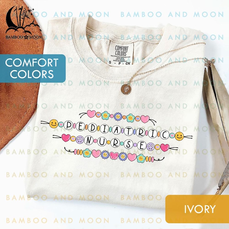 May include: Cream-coloured t-shirt featuring the text "PEDIATRIC NURSE" in a colourful, playful font. The design includes hearts, flowers, peace signs, and smiley faces. A gold necklace with a pendant rests on the shirt.