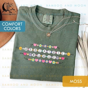 May include: A moss green Comfort Colors t-shirt featuring the words "PEDIATRIC NURSE" in a colourful, playful design. The design includes hearts, flowers, peace signs, and smiley faces. A silver necklace with a pendant is also visible.