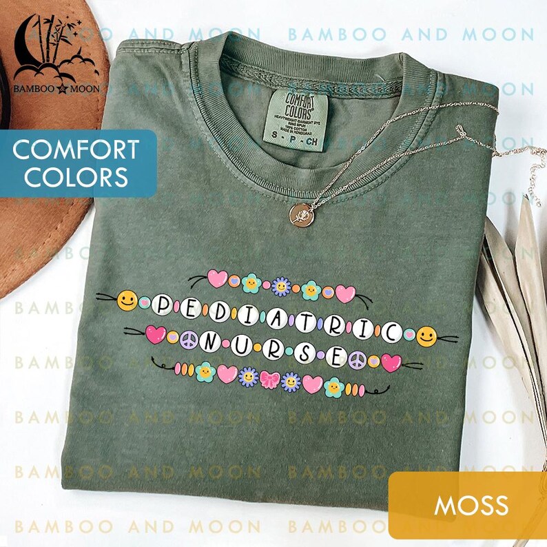 May include: A moss green Comfort Colors t-shirt featuring the words "PEDIATRIC NURSE" in a colourful, playful design. The design includes hearts, flowers, peace signs, and smiley faces. A silver necklace with a pendant is also visible.
