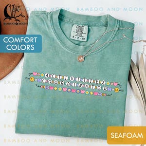 Könnte beinhalten: Seafoam-grünes Comfort Colors T-Shirt mit den Worten "ACTIVITIES COORDINATOR" in einem Perlendesign. Das Design umfasst Herzen, Blumen und Smiley-Gesichter. Eine silberne Halskette mit Anhänger liegt auf dem Shirt.